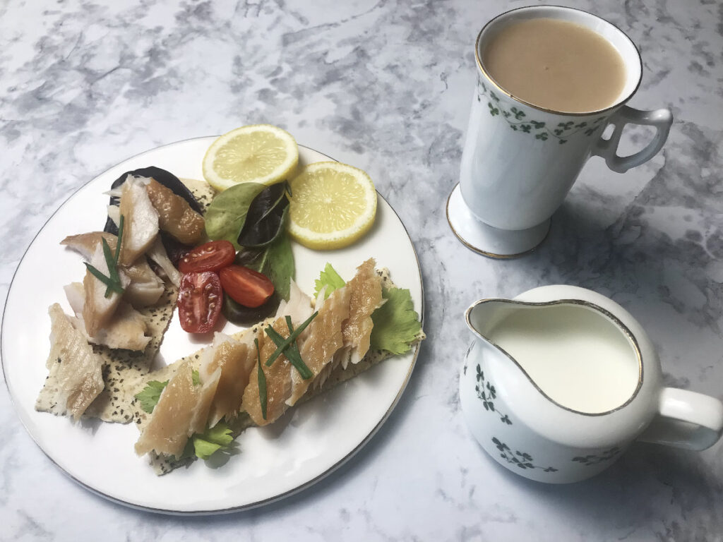 White plate on the left filled with seeded crackers topped with Smoked Trout garnished with chives, served with a side salad and some fresh lemon slices. To the right is a mug of tea and a milk jug filled with milk.