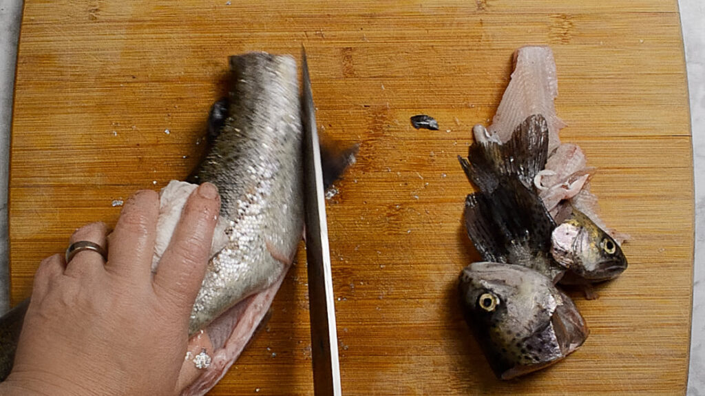 Wooden board with a rainbow trout on it, the head and tail are on the right of the board, and on the left is the body of the fish with the fins being sliced off. A hand is holding the fish and a knife is slicing through the fins.