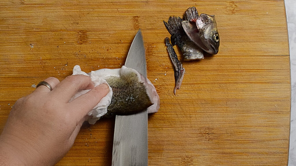 Wooden board with a rainbow trout on it, the head and tail are on the right of the board, and on the left is the body of the fish. The top filet of the Trout is being removed using a knife. A hand is holding the fish. 