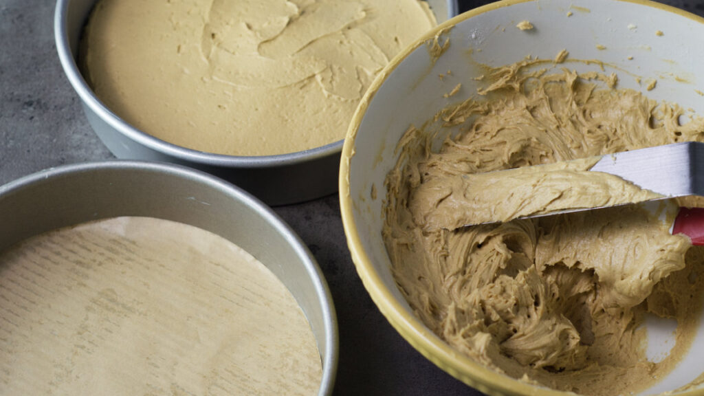 To the right is a bowl filled with coffee cake batter. In the bowl is a metal spatula knife. To the left are the 2 cake pans, one contains cake batter and the other is empty.