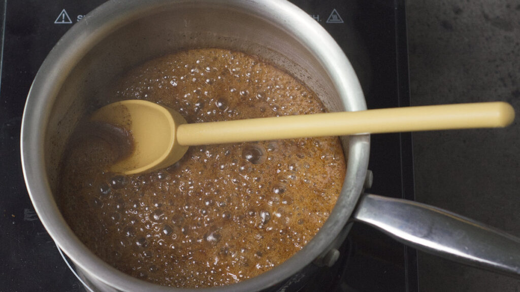 Pot on the burner containing foamy sugar. There is a wooden spoon in the pot.