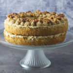 Coffee Walnut Sandwich showing the layers of sponge, buttercream and walnuts on top. The cake is on a glass cake stand.