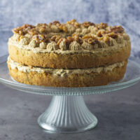 Coffee Walnut Sandwich showing the layers of sponge, buttercream and walnuts on top. The cake is on a glass cake stand.