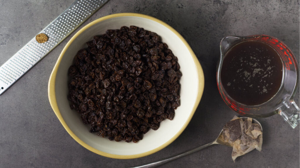 Bowl containing raisins and spices in the center. To the left is nutmeg resting on a grater. To the right is a jug of strong tea with spent teabags beside the jug.
