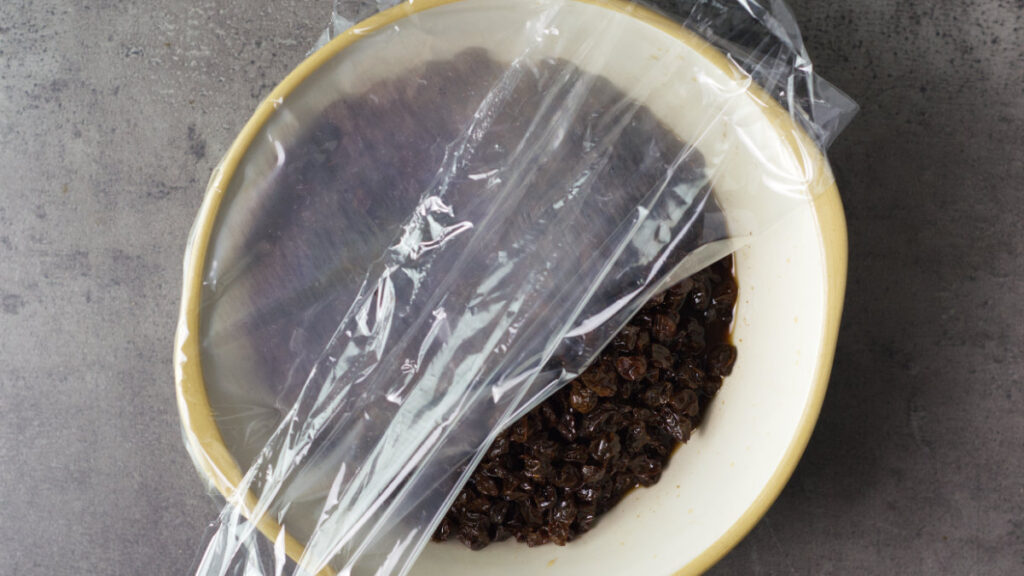 Bowl containing steeped raisins. The plastic wrap covering the bowl is partially removed.