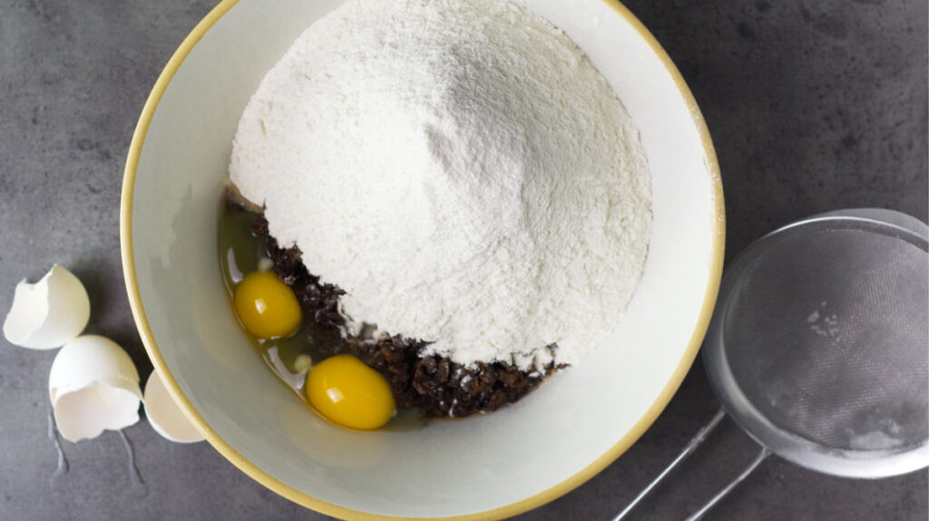 Bowl with steeped raisins, flour, eggs, baking powder and salt added. To the left are egg shells and to the right is the sieve.