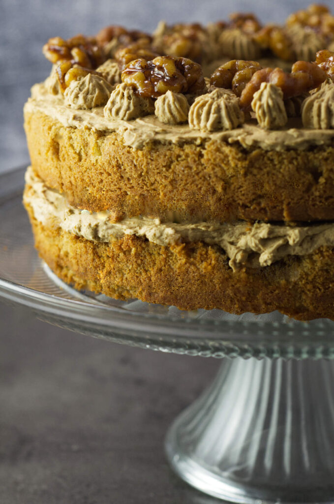 Side view of the Coffee Walnut Sandwich showing the layers of sponge, buttercream and walnuts on top. The cake is on a glass cake stand.
