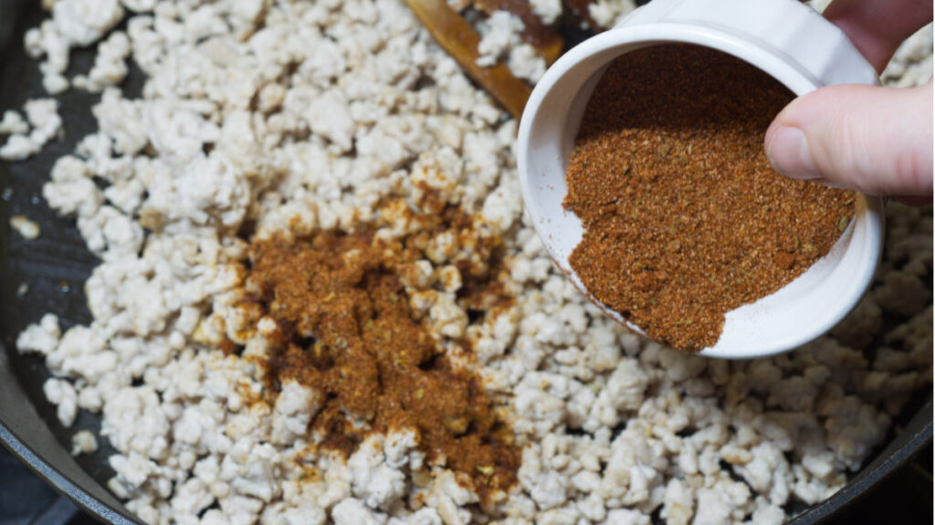 Black pan with cooked ground in it, chorizo seasoning is being added.