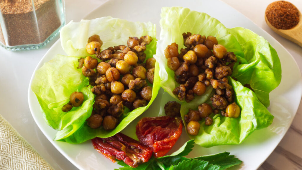White plate in the foreground with chorizo chickpeas lettuce cups on the plate with a tomato and parsley garnish. In the background is a jar of chorizo seasoning with a spoon of seasoning beside it.