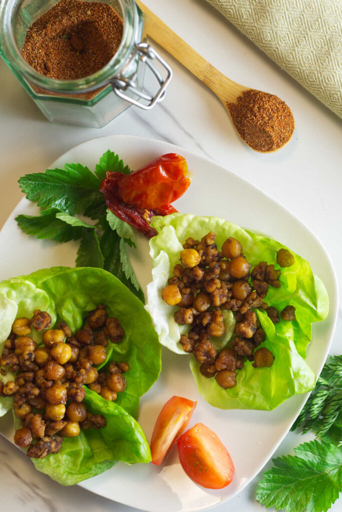 White plate in the foreground with chorizo chickpeas lettuce cups on the plate with a tomato and parsley garnish. In the background is a jar of chorizo seasoning with a spoon of seasoning beside it and a pale green napkin.