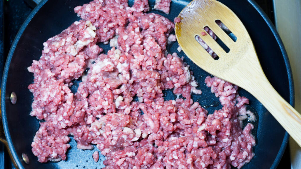 Black pan containing the ground lamb which is in the process of being cooked. A wooden spoon is resting in the pan.