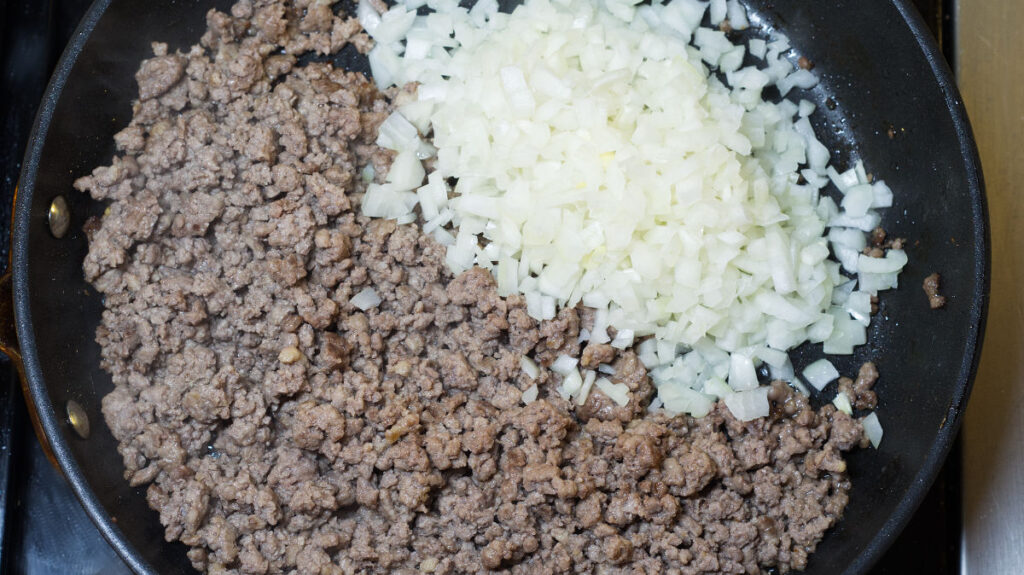 Black pan containing cooked ground lamb and chopped onion added.