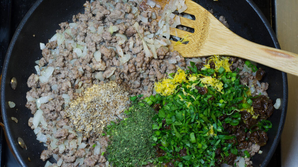Black pan with cooked lamb, diced onions and garlic in it. Chopped parsley, scallions, dried herbs, lemon zest  and spices have been added. A wooden spoon is resting in the pan.