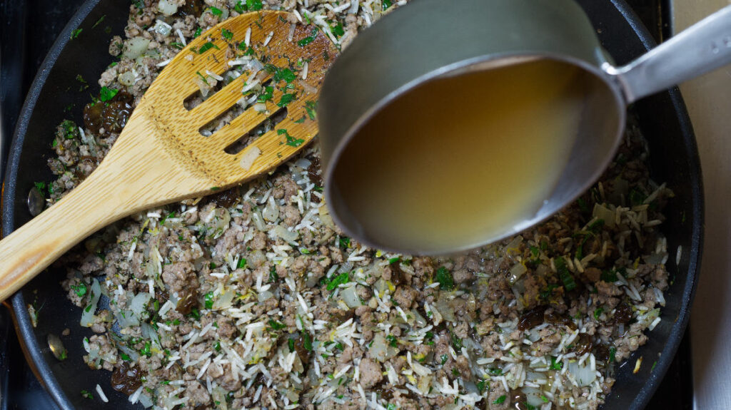 Black pan with cooked stuffing mixture in it. Chicken stock is added. A wooden spoon is resting in the pan.