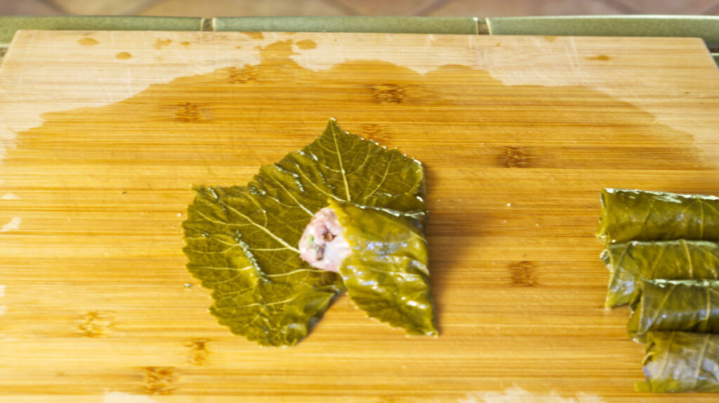 Wooden board with a grape leaf. On it is a stuffing sausage with the right side of the grape leaf folded over the stuffing. To the right are completed rolled up stuffed grape leaves.