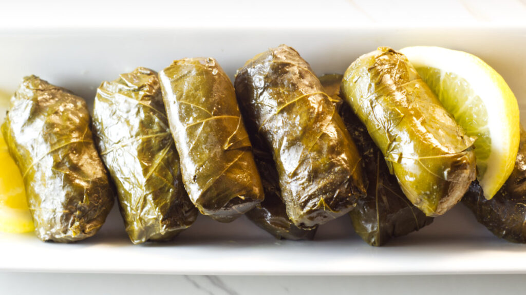 White rectangular plate with cooked stuffed grape leaves on it, garnished with lemon wedges.