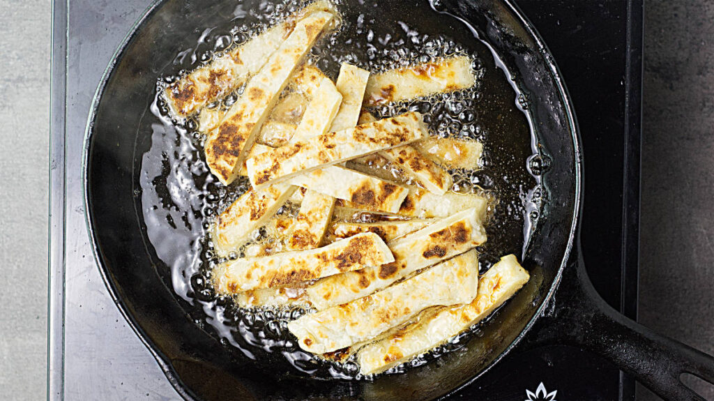 Black skillet with hot fat and sizzling potato farl strips. 