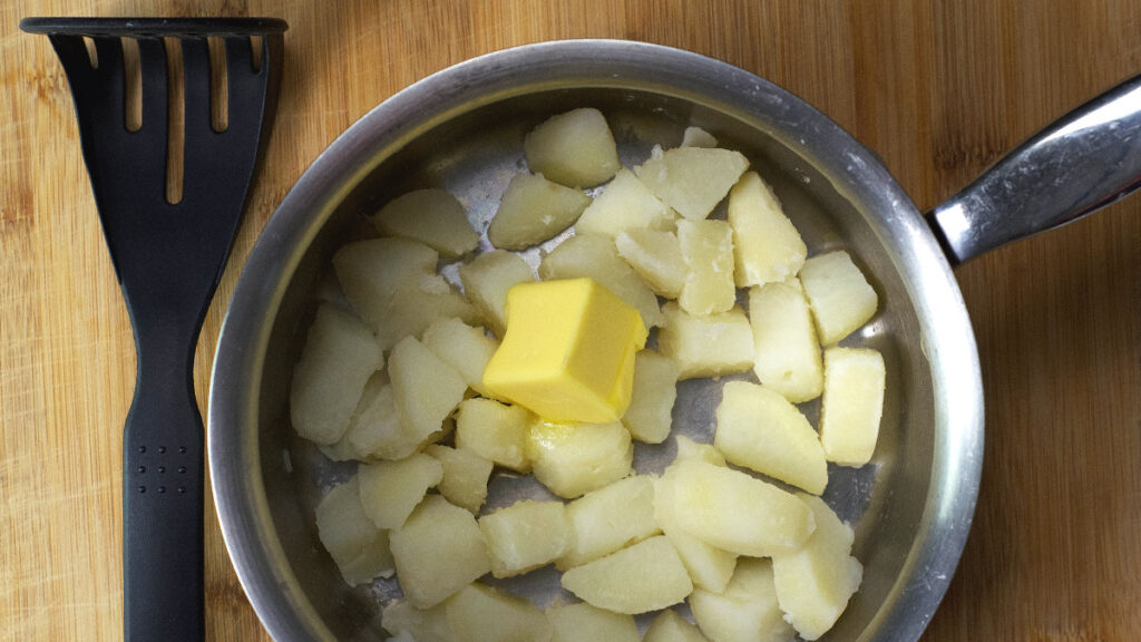 Pot containing cooked potatoes with a knob of butter in them and a potato masher resting on the board.
