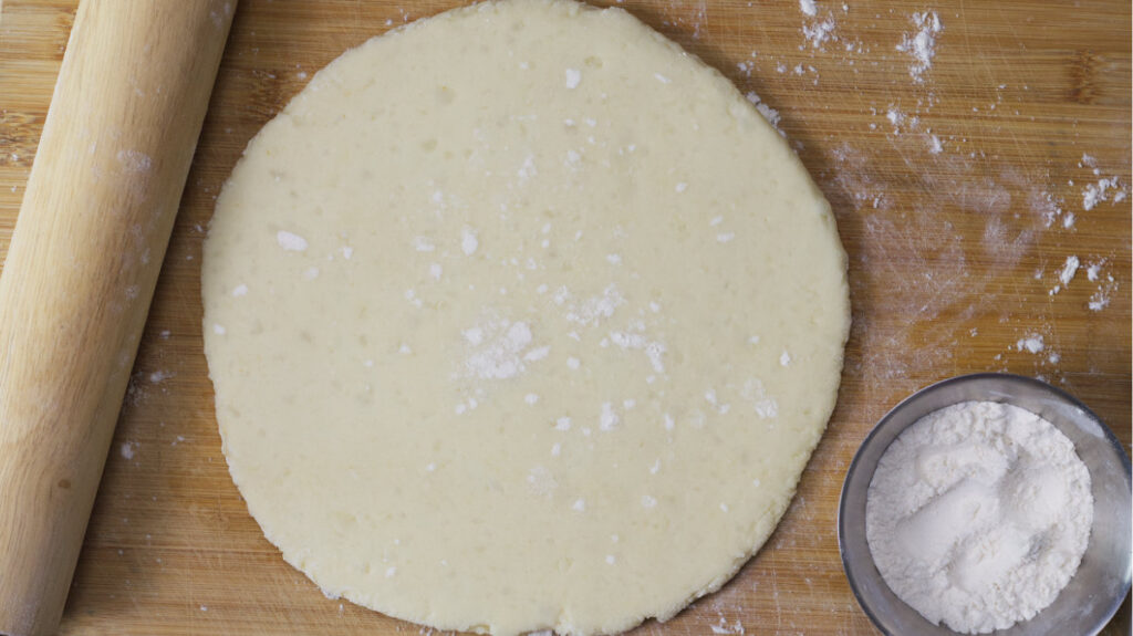 Dough is rolled out in a circle on the wooden board. Resting on the board is a wooden rolling pin and a small bowl of flour.