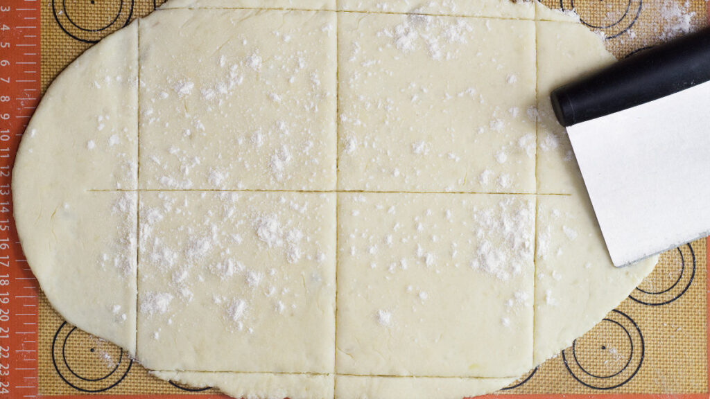 Uncooked potato dough rolled out and cut into squares. Pastry cutter is resting beside the rolled out dough.