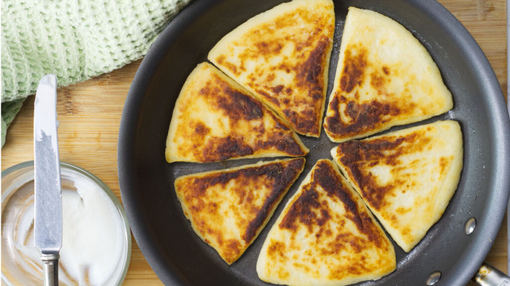 Skillet with cooked Potato Farls, to the side is a green drying cloth and a bowl of bacon fat with a knife resting on top.